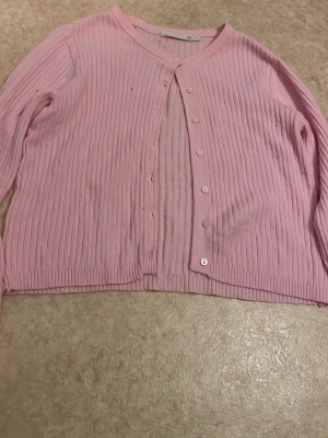 Ljusrosa ribbad kofta - Söt ljusrosa kofta med ribbad struktur och rund hals. Koftan har långa ärmar och knäppning framtill med matchande rosa knappar. Perfekt att slänga över en topp för en chill look.