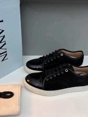 Svarta Lanvin sneakers i mocka och lack - Snygga svarta sneakers från Lanvin med ovandel i mocka och lackad tå. Skorna har vita sulor, svarta skosnören och metallfärgade öljetter. Klassisk låg modell som passar till det mesta och ger en clean look storlek 41 men sitter som 42 box och allt og finns kvitto finns ochså (ÄKTA)⚠️⚠️