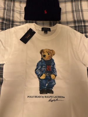 Vit Polo Bear t-shirt från Ralph Lauren Medium och mössa - Vit t-shirt från Ralph Lauren med ikoniska Polo Bear-trycket framtill. Björnen har på sig jeansjacka och jeans, och under motivet står det 'POLO BEAR by RALPH LAUREN'. Klassisk rund hals och korta ärmar. Perfekt statement-plagg för dig som gillar streetstyle.