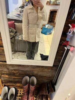 Vit trenchcoat med bälte - Stilren vit trenchcoat med bälte i midjan och stora fickor framtill från märket ICHI. Jackan har klassisk krage, knäppning med knappar och justerbara ärmslut. Perfekt för dig som vill ha en tidlös och snygg look. Den är perfekt för våren