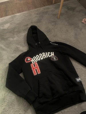Svart hoodie från Hoodrich - Snygg svart hoodie från Hoodrich med stor broderad H och vita samt röda detaljer. Hoodien har huva med dragsko, känguruficka och coola patches på bröstet. Perfekt för dig som gillar streetwear och vill sticka ut lite extra. Nytt pris var 750kr