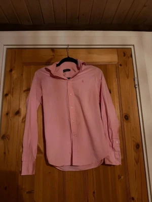 Rosa skjorta från Polo Ralph Lauren - Klassisk rosa skjorta från Polo Ralph Lauren med broderad logga på bröstet. Skjortan har lång ärm, knappar framtill och en tidlös krage. Perfekt för dig som gillar stilrena och snygga plagg.