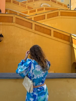 Blått Nelly Hypnotic Beach Set - Supersnyggt tvådelat set i blått mönster. Består av en långärmad croppad topp med knytdetalj framtill samt en matchande sarong-/omlottkjol som knyts i sidan. Perfekt till strand, semester, beach club eller sommarfest.