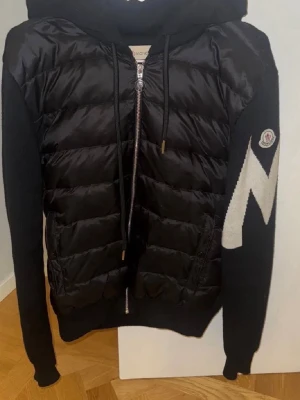 Moncler cardigan - Snygg svart moncler cardigan i storlek M med quiltad front och stickade ärmar. Jackan har dragkedja, huva med snörning och ett stor vitt M på ärmen samt Moncler logga. Perfekt för dig som vill ha en skön och trendig look. Svarar alla meddelanden direkt och fraktar alltid samma dag som köp:)