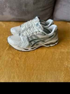 Vita sneakers från Asics - Säljer ett par vita Asics sneakers med mesh och syntetpaneler. Skorna har grå och silverfärgade detaljer samt klassiska Asics-loggan på sidan. Lågt skaft och snörning framtill. Perfekta för en sportig och trendig look.