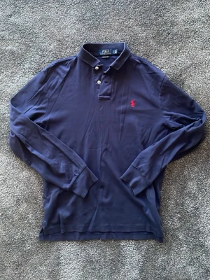 Marinblå långärmad piké från Polo Ralph Lauren - Säljer en marinblå långärmad pikétröja från Polo Ralph Lauren med klassisk krage, knappar och röd broderad logga på bröstet. Modellen är Custom Slim Fit och materialet är mjuk bomull. Perfekt för en clean och stilren look.