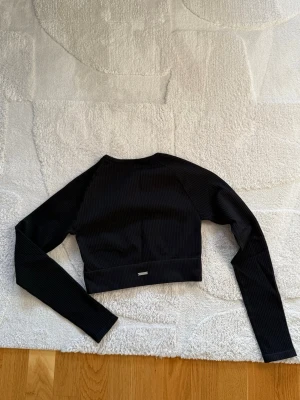 Svart ribbad croppad topp. Aim’n - Snygg svart croppad topp med långa ärmar och ribbad struktur. Perfekt för en trendig och sportig look. Liten metallbricka med logga baktill i midjan ger en extra detalj. Passar till både jeans och kjol.