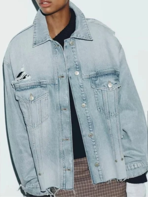 Oversized ljusblå jeansjacka - Trendig oversized jeansjacka i ljusblå denim med slitningar på axel och bröstficka. Klassisk krage, två bröstfickor med lock och knappar framtill. Perfekt för en avslappnad streetstyle-look.