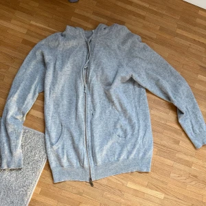 Ljusgrå cashmere  hoodie med dragkedja från Davida - Mysig ljusgrå hoodie från Davida med dragkedja framtill och två fickor. Tillverkad i mjukt material och har en klassisk huva. Perfekt för chill dagar eller när du vill ha en enkel och stilren look.