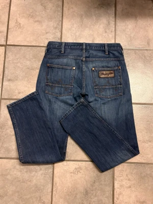Blå Wrangler jeans med raka ben - Säljer ett par klassiska blå jeans från Wrangler med raka ben och snygga detaljer på bakfickorna. Jeansen har fem fickor, bälteshällor och tydliga kontrastsömmar. Perfekta för en avslappnad och tidlös look.