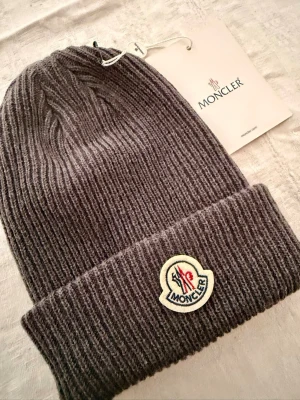 Ribbstickad Moncler mössa - Säljer en ribbstickad charcoalfärgad mössa från Moncler med uppvikt kant och klassisk Moncler-logga framtill. Mössan är tillverkad i mjukt material och har stilren, tidlös design. Oanvänd med patch.