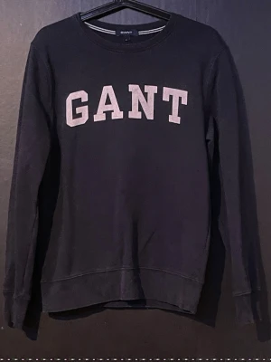 Mörkblå sweatshirt från GANT storlek s - Klassisk mörkblå sweatshirt från GANT med stora vita bokstäver på bröstet. Tröjan har rund halsringning, långa ärmar och ribbade muddar vid ärmslut och nederkant. Perfekt för en avslappnad och stilren look. Storlek s och har aldrig använt 