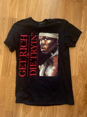 Svart 50 Cent t-shirt Get Rich or Die Tryin' - Svart t-shirt från 50 Cent med stort tryck av albumomslaget 'Get Rich or Die Tryin'' på framsidan. T-shirten är i 100% bomull och har klassisk passform med korta ärmar och rund hals. Perfekt för dig som gillar hiphop och streetwear.