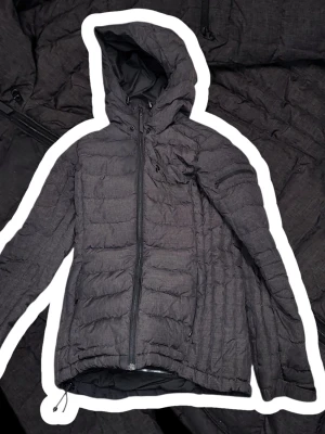 Peak Performance Jacka Dunn - Säljer en svart pufferjacka med huva och dragkedja framtill. Jackan har quiltad design, dragsko nertill och flera fickor med dragkedja. Perfekt för kalla dagar och har en stilren look som passar till det mesta. Materialet är syntet och jackan är lätt vadderad.