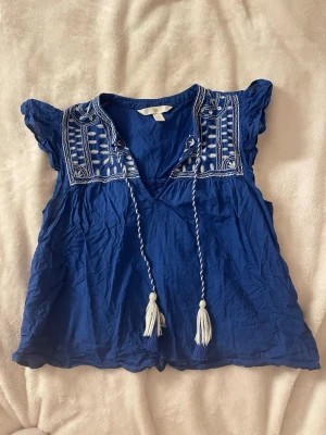 Blå blus med broderi från H&M - Snygg blå  blus från HM med vita broderier på axlarna och knytband med tofsar. Toppen har korta volangärmar och v-ringning. Den är ganska skrynklig men man kan säkert stryka den.