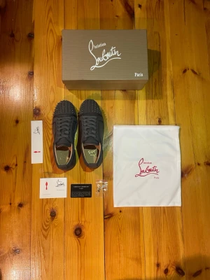 Grå Christian Louboutin sneakers med nitar - Grå sneakers från Christian Louboutin med ikonisk röd sula och coola nitar på tån. Skorna har snörning och är gjorda i mocka med läderfoder. Kommer med originalkartong, dustbag och certifikat. Perfekt för dig som vill sticka ut med lyxiga detaljer.