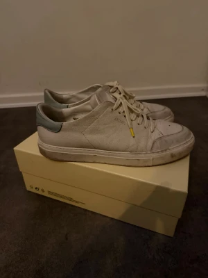 Vita Axel Arigato sneakers i läder - Snygga vita sneakers från Axel Arigato i läder med diskret logga på sidan. Skorna har klassisk låg profil, vita snören med gula detaljer och en grå detalj vid hälen. Perfekta för en clean och stilren look. INGA SKADOR BAGA LITE SMUTS, Är i jätte bra skick, 