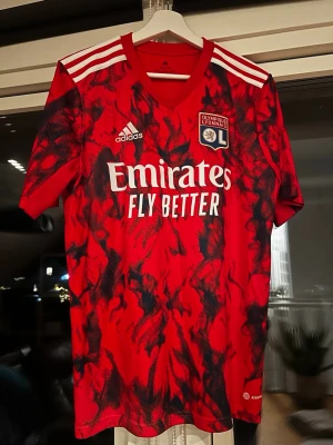 Olympique Lyonnais röd matchtröja Adidas - Säljer en röd och svart Olympique Lyonnais matchtröja från Adidas med vita detaljer och klubbmärke på bröstet. Tröjan har korta ärmar, V-ringning och är tillverkad i ett lätt, återvunnet funktionsmaterial. Snyggt mönster och Emirates Fly Better-tryck på framsidan.