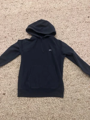 Navy hoodie från Stüssy - Snygg mörkblå hoodie från Stüssy med klassisk logga broderad på bröstet. Hoodien har en stor känguruficka framtill, dragsko i huvan och är tillverkad i mjukt bomullsmaterial. Perfekt för en avslappnad streetwear-look.