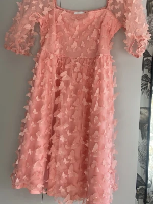 Rosa kortklänning med 3D fjärilar - Supersöt rosa kortklänning med voluminösa, halvtransparenta ärmar och 3D-applikationer i form av fjärilar över hela klänningen. Klänningen har en luftig passform och är perfekt för dig som vill sticka ut. Materialet känns lätt och har en mesh-liknande struktur.