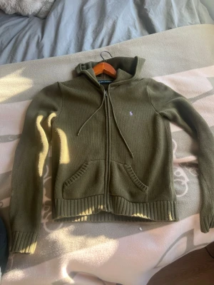 Grön stickad hoodie från Ralph Lauren - Säljer en  stickad hoodie från Ralph Lauren med dragkedja framtill och två fickor. Tröjan har huva med dragsko och den klassiska lilla loggan broderad på bröstet. Perfekt för en chill och stilren look. Storlek L men passar S 