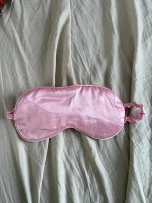 Rosa sovmask i satin - Snygg och mjuk sovmask i glansigt rosa satinmaterial. Masken har en elastisk rem med rynkdetalj för bekväm passform. Perfekt för att blockera ljus när du vill vila eller sova extra gott. Den här en fläck på sig, borde gå bort i tvätten