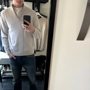 Beige halvzip tröja - Snygg beige tröja med halv dragkedja och hög krage. Tröjan har en avslappnad passform och ribbade muddar vid ärmslut och nederkant. Perfekt att bära över en t-shirt eller skjorta för en clean och stilren look. 