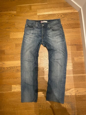 Levi's 511 blå jeans herr - Säljer ett par klassiska Levi's 511 jeans i blå denim med snyggt slitna detaljer och raka ben. Jeansen har fem fickor, knappgylf och den ikoniska röda Levi's-lappen bak. Perfekta för en avslappnad och stilren look. Jeansen är i längd 34 men uppsydda så de passar 30 i längd 