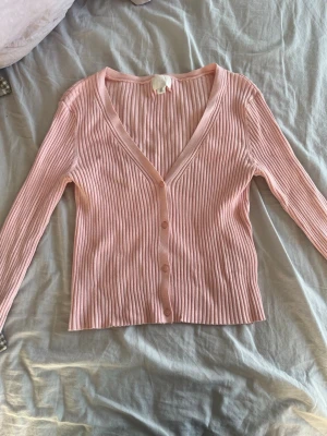 Ljusrosa ribbad kofta från H&M med knappar - Söt ljusrosa kofta från H&M med ribbad struktur och v-ringning. Koftan har långa ärmar och knäpps med matchande knappar framtill. Perfekt att slänga över en topp för en chill look.