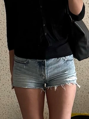 Ljusblå jeansshorts med fransig kant - Säljer ett par ljusblå denimshorts med fransig, rå kant och klassisk femficksdesign. Shortsen har låg midja och en avslappnad passform, perfekta för varma dagar. Snygga att matcha med en enkel topp eller skjorta. Shortsen är från levis