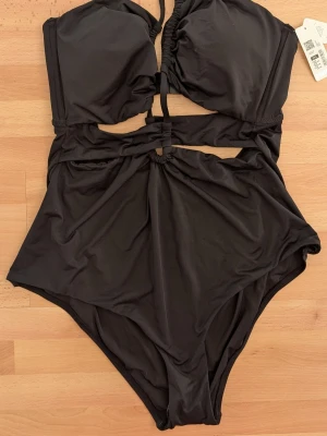 Cubus Svart baddräkt, Baddräkt i ett stycke  - Trendig svart baddräkt med snygga cut-outs och rynkade detaljer framtill. Baddräkten har vadderade kupor och smala axelband som knyts i nacken. Tillverkad i stretchigt material som passar perfekt för stranden eller poolen.