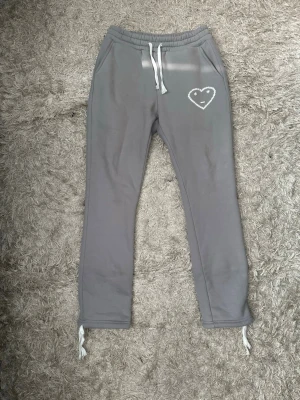 Gråa mjukisbyxor från carsicko - Säljer ett par grå mjukisbyxor från carsicko med vita dragsnören i midjan och vid bensluten. Byxorna har fickor fram och en söt vit hjärtdetalj på ena sidan. Materialet är mjukt och skönt, perfekt för chill dagar. Bakficka finns också. Passar perfket med den matchande hoodien. Hela setet/tracksuiten finns i min profil som en egen annons.