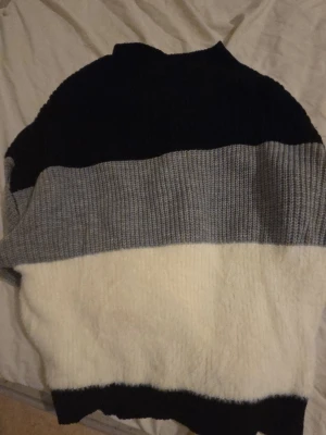 Randig stickad tröja i svart, grått och vitt - Mysig stickad tröja med breda blockränder i svart, grått och vitt. Tröjan har en avslappnad passform och ribbstickade detaljer vid hals, ärmslut och nederkant. Perfekt för kalla dagar och enkel att matcha med jeans eller kjol.