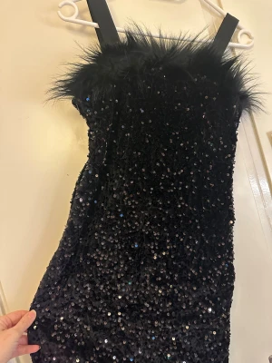 Svart paljettklänning med fuskpäls - Supersnygg svart kort klänning täckt av glittriga paljetter och med breda axelband. Överdelen har en fluffig kant i fuskpäls som ger en extra cool touch. Perfekt för dig som vill sticka ut och glänsa på festen!
