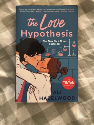 The Love Hypothesis av Ali Hazelwood - En romantisk och humoristisk roman på engelska, 'The Love Hypothesis', med ett blått och rosa omslag som visar två tecknade personer i labbmiljö. Boken är en TikTok-sensation och handlar om kärlek, vetenskap och fejkat förhållande.