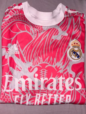 Real Madrid matchtröja rosa - Snygg rosa Real Madrid matchtröja med vitt drakmönster, klubbmärke och sponsortryck. På ryggen står det Ronaldo och nummer 7. Tillverkad i lätt och ventilerande material. Passar mig som har S/XS