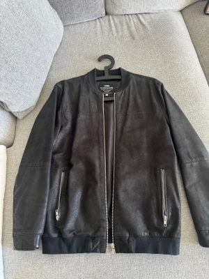 Svart skinnjacka från Jack & Jones - Snygg svart skinnjacka från Jack & Jones med raka linjer och minimalistisk design. Jackan har två framfickor med dragkedja och ribbade muddar vid ärmslut och nederkant. Perfekt för en stilren look.