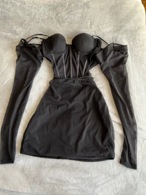 Svart kort klänning med puffärmar - Trendig svart kort klänning med markerad byst, puffiga långärmar och smala axelband. Klänningen har en kroppsnära passform och detaljerade sömmar framtill som ger en snygg siluett. Perfekt för dig som vill sticka ut med en edgy look.     Stl: Uk-6 US-2