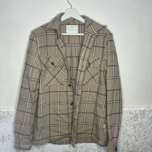 Beige rutig overshirt från Jackn Jones - Stilren beige overshirt från H&M med svart rutmönster. Två bröstfickor med knappar och klassisk krage. Perfekt lager-på-lager-plagg med avslappnad passform och mjukt material. Passar till jeans eller chinos för en trendig look.