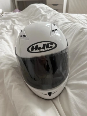 Vit integralhjälm från HJC - Säljer en vit integralhjälm från HJC med svart visir och tydlig logga framtill. Hjälmen har ventilationsöppningar och en aerodynamisk form. Insidan är vadderad för extra komfort och visiret kan fällas upp. Perfekt för motorcykel eller moped.