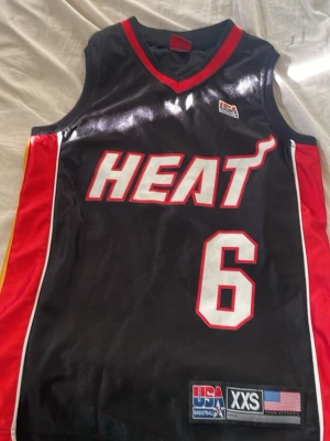 Miami Heat basketlinne James #6 - Snyggt svart basketlinne från Miami Heat med röd och gul detalj på sidorna. Nummer 6 och namnet James på ryggen. Tillverkat i lätt och ventilerande material, perfekt för basket eller streetwear.