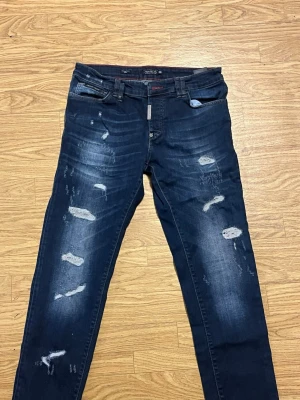Mörkblå slitna jeans från Philipp Plein - Säljer ett par mörkblå jeans från Philipp Plein med slitningar och distressed detaljer framtill. Jeansen har raka ben, klassisk femficksmodell och en patch med PP-logga på bakfickan. Materialet är denim i bomull med stretch för extra komfort.