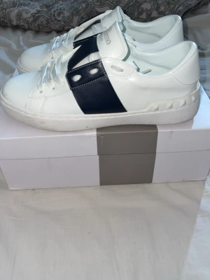 Valentino vita sneakers med svart detalj - Snygga vita sneakers från Valentino Garavani med bred svart läderdetalj och nitar på sidan. Klassisk låg modell med rund tå och vita skosnören. Tillverkade i slätt läder för en clean och exklusiv look. Perfekta för dig som vill ha något stilrent men ändå sticker ut.