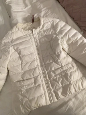 Vit pufferjacka från Lindex  - Säljer en stilren vit pufferjacka från Lindex. Jackan har en klassisk quiltad design, rund halsringning och dragkedja framtill. Perfekt för kalla vinterdagar och lätt att matcha med olika outfits. Materialet är lätt och värmande.