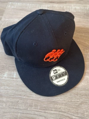 Svart snapback-keps Monkey Shoulder - Snygg svart snapback-keps från New Era 9FIFTY med broderad orange text 'Monkey Shoulder' på baksidan och en cool orange logga framtill. Klassisk platt skärm och justerbar passform. Tillverkad i slitstark bomull.