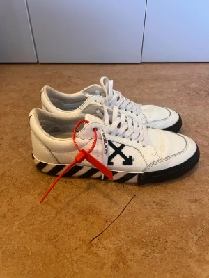 Off-White vita sneakers med ränder - Säljer ett par vita Off-White sneakers i läder med svarta pilar på sidan och svartvita ränder på sulan. Skorna har vita skosnören med text och en röd tag som signaturdetalj. Klassisk low-top modell med chunky sula och streetwear-vibe.