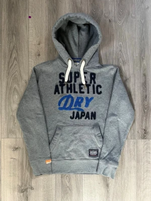 vintage Superdry hoodie med tryck - Snygg grå hoodie från Superdry med broderat texttryck i svart och blått på bröstet🤩 Passar perfekt för nån i S och inga fel😇 Priset är ej hugget i sten🙌🏻