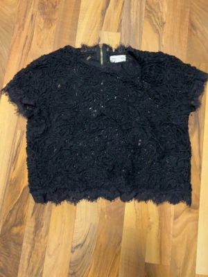 Svart spetsblus med dragkedja - Kort svart blus från Gina Tricot med vackra spetsdetaljer och blommönster. Toppen har korta ärmar, rund halsringning och en synlig dragkedja bak. Perfekt för dig som gillar en unik och trendig look.