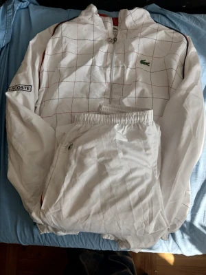 Vit träningsdress från Lacoste - Säljer en vit träningsdress från Lacoste med diskret rutmönster i rött och blått. Jackan har dragkedja, hög krage och logga på bröstet samt ärmen. Byxorna har resår i midjan och sidoficka med dragkedja. Perfekt för sport eller chill.