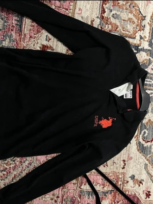 Svart långärmad piké från USPA - Svart långärmad pikétröja från USPA med röd logga och broderad häst på bröstet. Klassisk krage med knappar och kontrastfärgad insida i rött. Perfekt för en sportig och stilren look.